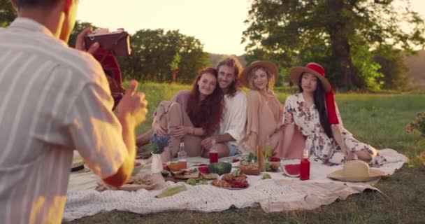 El hombre joven tomando la foto en la cámara vieja de las personas alegres diversas en el parque. Millennail amigos sonriendo y posando mientras hacen un picnic y se sientan en la hierba. Concepto de momentos felices. — Vídeo de stock