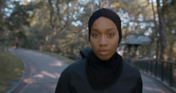 Vista da vicino della giovane donna musulmana in tuta sportiva che corre nel parco. Persona femminile energica in hijab che fa jogging mentre guarda alla telecamera. Concetto di stile di vita sano, sport. All'aperto. — Video Stock