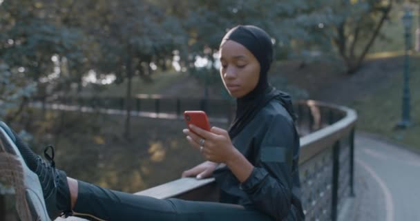 Muslinská žena se zahřívá a používá smartphone v parku. Žena v hidžábu zvedá hlavu a usmívá se při protahování při rolování obrazovky telefonu venku. Přiblížit. Pojetí sportu. — Stock video