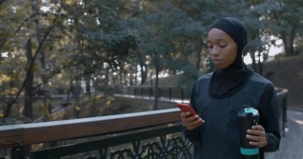 쾌활 한 여성이 스마트폰을 사용하고 스포츠용 병에서 물을 마시고 있습니다. 히잡을 끼고 공원을 걷고 있는 여성이 소셜 미디어에서 수다를 떨고 있는 모습이 눈에 띈다. 건강 한 생활 방식에 대한 개념. — 비디오