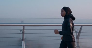 Köprüde koşan, spor takım elbiseli, güzel görünümlü müslüman bir kadının yan görüntüsü. Başörtüsü ve koşu giymiş profesyonel bir sporcu. Sağlıklı yaşam tarzı kavramı, spor. Açık havada..