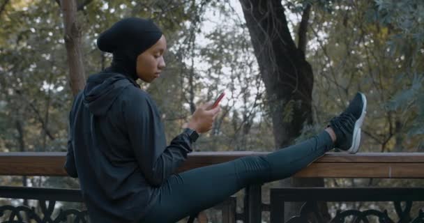 젊은 미국 여성들이 공원에서 스마트폰을 사용하면서 따뜻 해 지고 있습니다. 히잡을 끼고 밖에서 핸드폰 화면을 스크롤하고 있는 여자. 건강 한 생활 방식, 스포츠의 개념. — 비디오