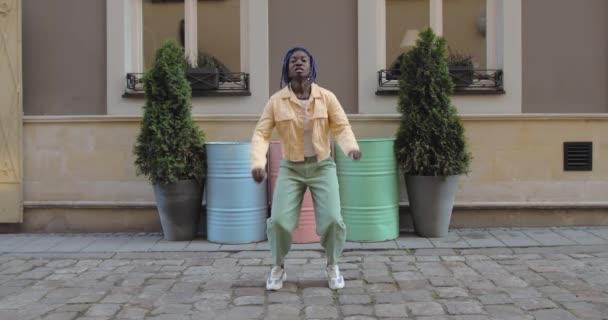 Female professional dancer moving in rhytm and smiling at city street. Afro american cheerful woman dancing while standing outdoors. Concept of good mood. Colourful barrels at background. — Stock Video