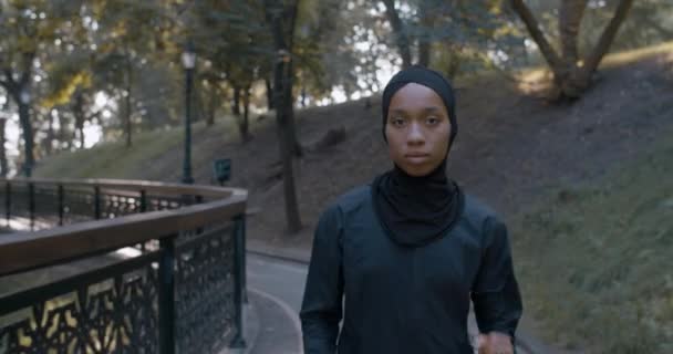 Вид спереду молода серйозна мусульманка, яка дивиться на камеру і бігає в парку. Приваблива жінка в хіджабі і спортивний костюм бігу. Концепція здорового способу життя, спорту. На відкритому повітрі . — стокове відео