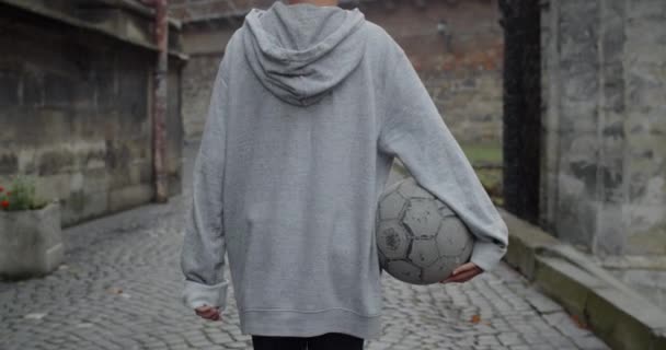 Back side view of teenager boy in hoodie carrying old football ball while walking at city street.Concept of sport, dreams and lifestyle.Outdoors. — Stock Video