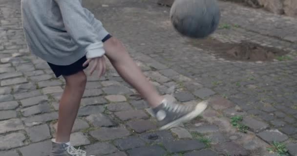 Vista de abajo hacia arriba de un niño adolescente con capucha pateando la pelota con los pies. Joven futbolista practicando trucos en la vieja calle de la ciudad. Concepto de deporte y estilo de vida. — Vídeos de Stock