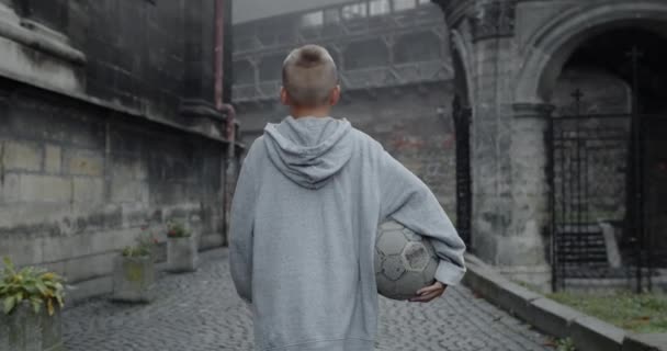 Beskär syn på tonåring kille i casual kläder som håller fotboll. Baksidan av pojke promenader på gamla europeiska gatan. Begreppet idrott, fritid, livsstil, barndom. Utomhus. — Stockvideo