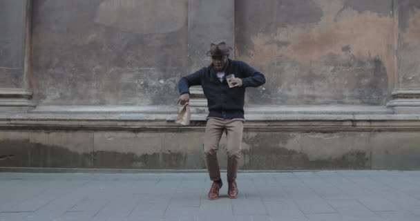 Guapo afroamericano con sombrero bailando mientras está parado en la vieja calle de la ciudad. Hombre positivo en ropa elegante que se mueve en rima mientras sostiene la taza de café de papel y la bolsa de artesanía. Al aire libre. — Vídeos de Stock