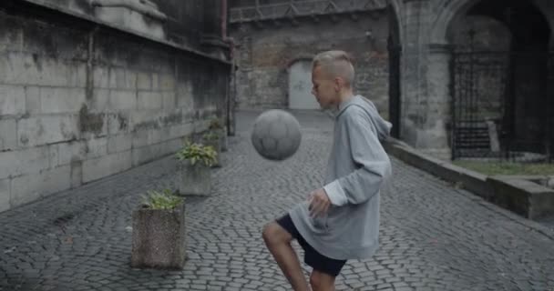 Vista lateral del jugador de fútbol adolescente niño pateando pelota de fútbol con rodilla. Chico con talento practicando trucos en la vieja calle de la ciudad. Concepto de deporte y estilo de vida. Persona en bicicleta en el fondo. — Vídeos de Stock