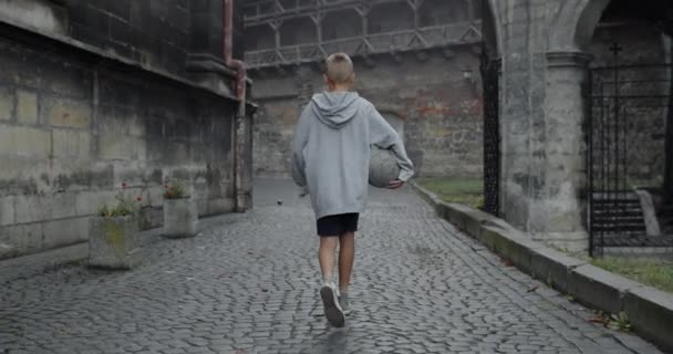 Visão traseira de criança adolescente em roupas casuais segurando bola de futebol. Jogador de futebol menino andando na velha rua da cidade europeia. Conceito de esporte, lazer, estilo de vida, infância. Ao ar livre. — Vídeo de Stock