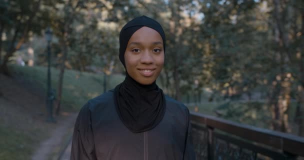 Porträtt av glada muslimska kvinna vrida huvudet och titta på kameran. Närbild av afro amerikanska kvinnlig person i hijab ler när du står i parken. Begreppet hälsosam livsstil. — Stockvideo