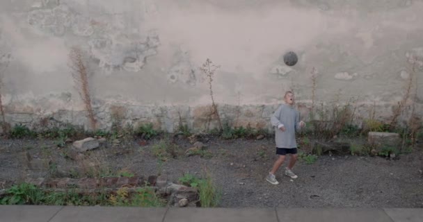 Młody dzieciak w kapturze odbijający piłkę nożną z głową na zewnątrz. Nastolatek trenujący sztuczki piłkarskie. Pojęcie stylu życia, dzieciństwa, sportu. — Wideo stockowe