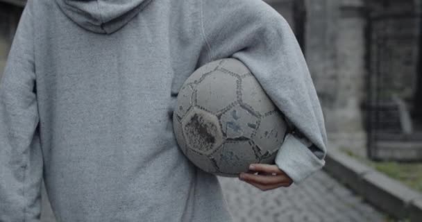 Baksidan av person i huvtröja bär gammal fotboll när du går på stadens gata.Begreppet sport, drömmar och livsstil.Utomhus. — Stockvideo