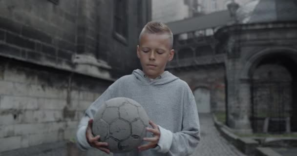 Portret uroczego nastolatka łapiącego i trzymającego piłkę nożną i patrzącego w kamerę. Widok ziarna białego chłopca pozowanie podczas stania na ulicy starego miasta. Pojęcie sportu. — Wideo stockowe