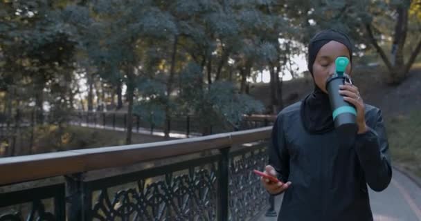 スポーツボトルからスマートフォンや飲料水を使用して若いイスラム教徒の女性。公園を歩いている間にソーシャルメディアをスクロールヒジャーブの女性の作物ビュー。健康的なライフスタイルの概念. — ストック動画