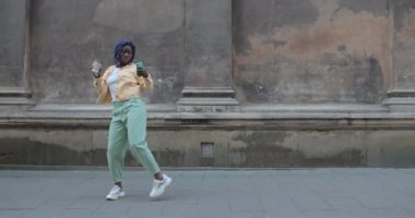 Eski şehir caddesinde akıllı telefon kullanırken dans eden duygusal Afro-Amerikan kadın. Milenyum kadını elinde kağıt kahve fincanı tutarak ritimle hareket ediyor. İyi ruh hali kavramı.