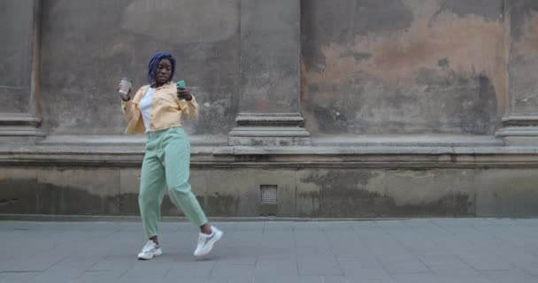 Emocional mujer afroamericana bailando mientras usa el teléfono inteligente en la vieja calle de la ciudad. Persona femenina milenaria sosteniendo taza de papel de café y moviéndose en rima. Concepto de buen humor. — Vídeos de Stock