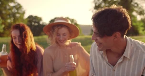 Oříznout pohled na tisícileté veselé lidi, kteří se smějí, zatímco drží skleněné láhve s pitím. Šťastní mladí lidé baví spolu a relaxaci při sezení na trávě v parku. — Stock video