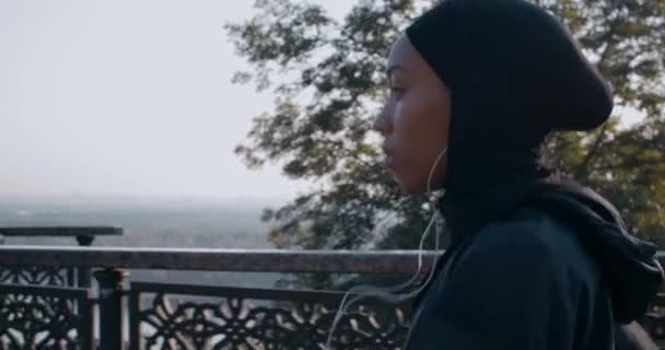 Nahaufnahme einer jungen muslimischen Frau mit Kopfhörern, die auf einer Brücke läuft. Seitenansicht einer weiblichen Person im Hijab, die beim Joggen Musik hört. Konzept eines gesunden Lebensstils. Draußen. — Stockvideo