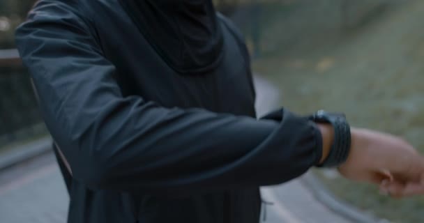 Vista de la cosecha de mujer desconocida en traje deportivo tocando la pantalla del reloj inteligente. Persona femenina en hijab comprobando gadget mientras está de pie en el parque. Concepto de estilo de vida saludable, deporte. — Vídeos de Stock