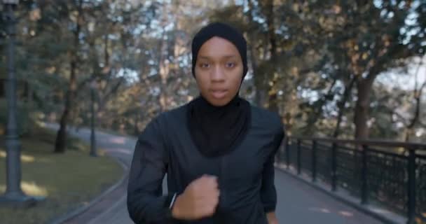 Vooraanzicht van de jonge mooie moslim vrouw die in het park rent. Energieke vrouwelijke persoon in hijab en sportpak joggen terwijl op zoek naar camera. Concept van gezonde levensstijl, sport. Buiten. — Stockvideo