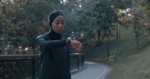 Vista de colheita da jovem mulher muçulmana em terno esportivo tocando exibição de relógio inteligente. Pessoa feminina em hijab olhando e verificando gadget inpark. Conceito de estilo de vida saudável. — Vídeo de Stock