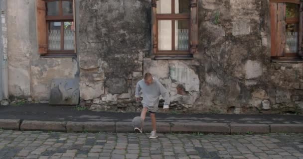 Nastolatek kopie piłkę nożną stopami. Młody piłkarz ćwiczący sztuczki na starej ulicy. Stara, zniszczona fasada budynku w tle. Pojęcie sportu i stylu życia. — Wideo stockowe