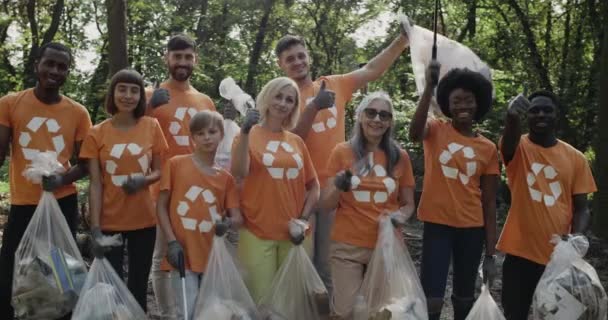 Veselí dobrovolníci v tričkách se symbolem recyklace se usmívají a ukazují palec nahoru. Skupina různých lidí, kteří se dívají na kameru, zatímco drží pytle s odpadky a stojí v parku. — Stock video