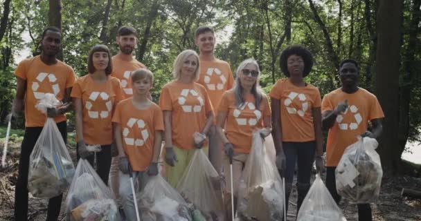 Volontaires multiethniques en t-shirts avec symbole de recyclage souriant et debout dans le parc. Groupe de personnes diverses tenant des sacs poubelle remplis de déchets et montrant pouce vers le haut tout en regardant à la caméra. — Video