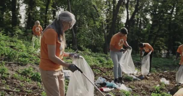 Mature lady using trash picker and holding bin bag while teen boy putting there plastic bottle. Diverse volunteers in t shirts with recycling symbol cleaning public park. Team work. — 图库视频影像