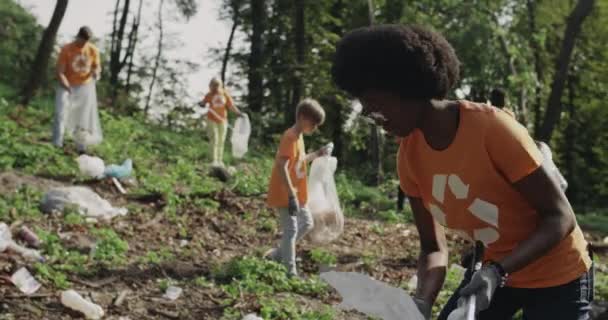Genç Afro-Amerikalı kadın çöp toplayıp çöp torbasına koyuyor. Kamu parkını temizleyen tişörtlü, geri dönüşüm tabelası olan bir grup çok ırklı gönüllü. Temizlik kavramı. — Stok video