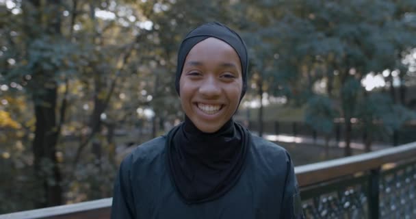 Вид на урожай красивой мусульманки, смеющейся и смотрящей в камеру. Портрет молодой веселой женщины в хиджабе, стоящей на улице. Концепция образа жизни, спорта. На открытом воздухе. — стоковое видео