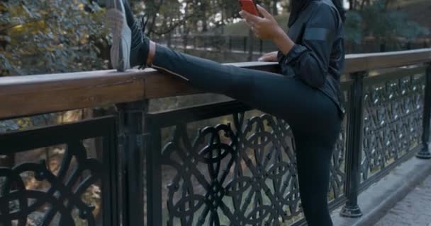 Ευτυχισμένη γυναίκα μουσελίνα προθέρμανση και χρήση smartphone στο πάρκο. Γυναίκα σε hijab χαμογελώντας και τεντώνοντας, ενώ κύλιση οθόνη του τηλεφώνου σε εξωτερικούς χώρους. Έννοια του υγιεινού τρόπου ζωής, αθλητισμός. — Αρχείο Βίντεο