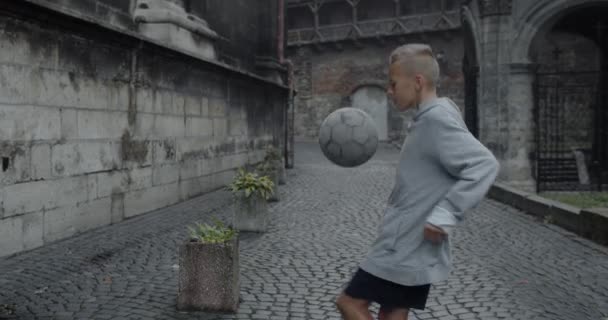 Side view of male kid soccer player kicking football ball with knee and head. Young boy practicing tricks at old city street. Concept of sport and lifestyle. — Stock Video