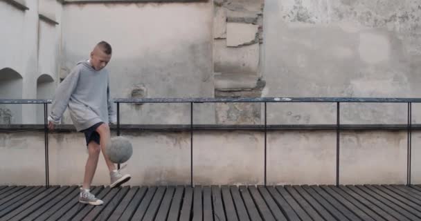 Young boy in hoodie bouncing football ball with leg at old city building background.Teenager warming up and playing football. Concept of lifestyle, childhood, sport. — Vídeo de Stock