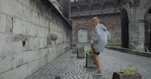 Side view of male kid soccer player kicking football ball with leg to wall . Young talented boy practicing tricks at old city street. Concept of sport and lifestyle. — Stockvideo