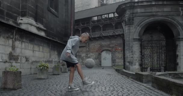 Een capabele tiener die voetbal schopt met voeten. Jonge getalenteerde jongen oefent trucs op de oude Europese stad straat. Begrip sport, talent en levensstijl. — Stockvideo