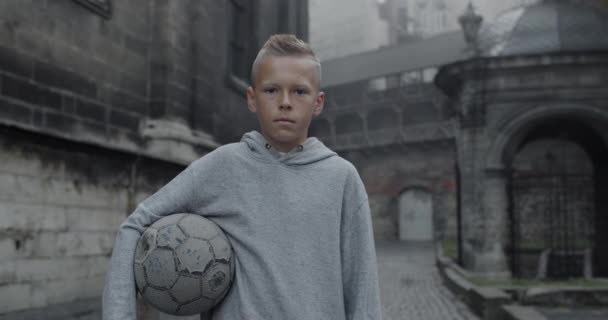Portrait of teenager kid holding football ball while looking and posing to camera. Crop view of young boy standing at old city street. Concept of sport. — Stockvideo