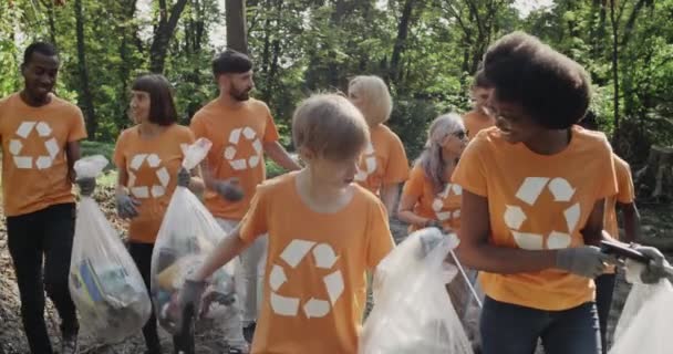 ゴミ箱袋を持っている多民族のエコ活動家やコミュニケーション。森林清掃後のシンボルウォーキングとトークをリサイクルしたTシャツの多様な人々のグループ. — ストック動画