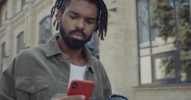 Siyah saçlı, kağıt kahve fincanı taşıyan ve internette gezinen yakışıklı Afro-Amerikan 'ı yakından izleyin. Genç sakallı adam şehir caddesinde yürürken akıllı telefon kullanıyor..