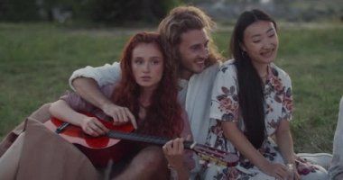 Başka bir kadın gitar çalarken genç bir adam Asyalı kadınla konuşuyor ve gülüyor. Neşeli gençler parkta çimenlerde iyi vakit geçiriyorlar..
