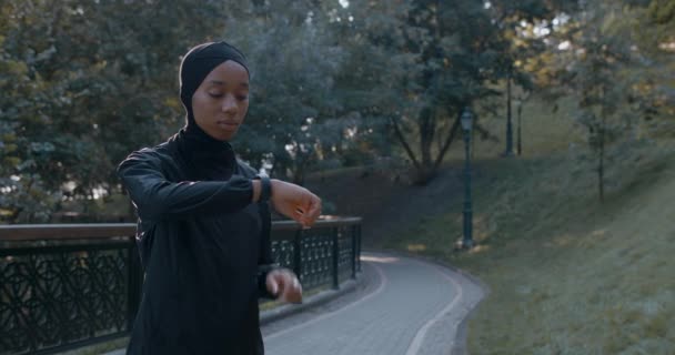 Vista de la cosecha de la mujer musulmana en traje deportivo tocando la pantalla del reloj inteligente. Joven mujer en hijab sonriendo y comenzó a correr en el parque después de comprobar el contenido del gadget. Concepto de deporte. — Vídeos de Stock