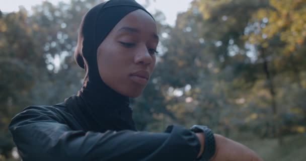 Vista de perto da mulher muçulmana em terno esportivo tocando exibição de relógio inteligente. Jovem mulher no hijab começou a correr no parque depois de usar o gadget. Conceito de esporte e estilo de vida saudável. — Vídeo de Stock