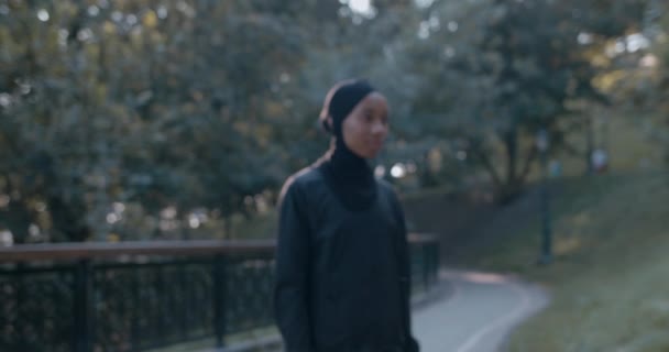 Schnittansicht einer fröhlichen muslimischen Frau im Sportanzug, die den Kopf dreht und in die Kamera blickt. Afroamerikanische Frau im Hidschab lächelt, während sie im Park steht. Vergrößern. — Stockvideo