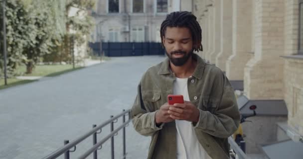 Άποψη των νέων μικτή φυλή άνθρωπος με dreadlocks περιήγηση στο διαδίκτυο, ενώ το περπάτημα στο δρόμο της πόλης. Γενειάδα χαρούμενος τύπος πληκτρολογώντας μήνυμα και χαμογελώντας κατά τη χρήση smartphone. — Αρχείο Βίντεο