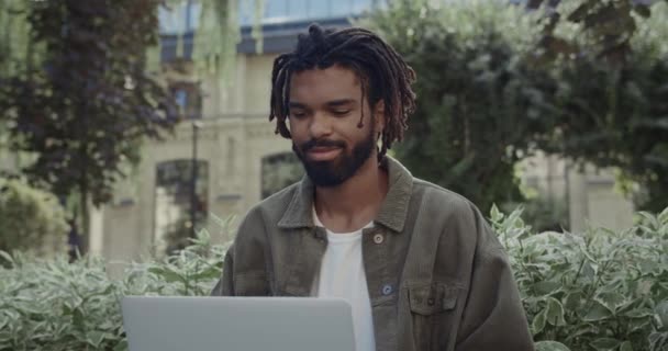 Schnittansicht von fröhlichen Kerl mit Dreadlocks mit Laptop im Park sitzend und lächelnd. Bärtiger gutaussehender afroamerikanischer Mann in lässiger Kleidung bei der Arbeit im Freien. — Stockvideo