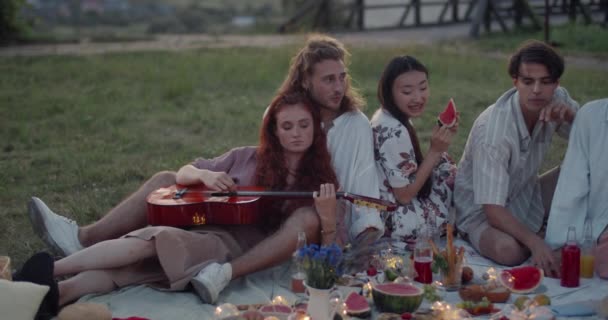 Jóvenes diversos hablando mientras están sentados en la hierba y haciendo picnic. Mujer atractiva tocando en la guitarra mientras se divierten juntos con amigos. Concepto de amistad y positivo. — Vídeo de stock