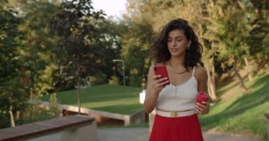 Sosyal medyada sohbet eden ve kahve fincanı taşıyan güzel İspanyol bir kadın. Mutlu esmer kadın internette geziniyor ve şehir parkında yürürken ekrana dokunuyor..