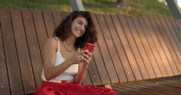 Обрезание зрения счастливой женщины с помощью смартфона во время чата в социальных сетях. Привлекательная женщина печатает сообщения, сидя на скамейке в парке. Концепция коммуникации. — стоковое видео