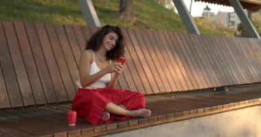 Sosyal medyada sohbet ederken akıllı telefon kullanan yalınayak neşeli bir kadın. Güzel bir kadın bankta oturup parkta kahve fren yaparken daktilo kullanıyor. İletişim kavramı.